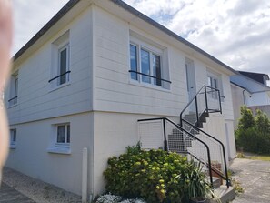 Ein zweistöckiges Gebäude mit weißer Fassade, einer Außentreppe und einem kleinen Gartenbereich mit Pflanzen.