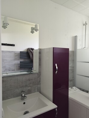 Een badkamer met een wit wastafel, een spiegel, een handdoekhouder, een paars kastje en een douchehoek.