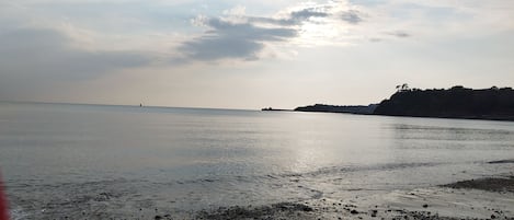 Uma praia com águas calmas, pedras e formações terrestres ao longe, sob um céu parcialmente nublado.