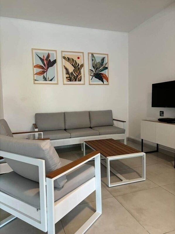 A modern living room with a grey sofa, a white armchair, and a wooden coffee table. There are three framed botanical prints on the wall.