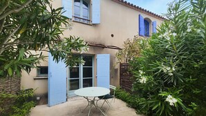 A patio with a table and chair, surrounded by lush greenery and a building with blue shutters.