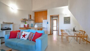 A living room with a blue sofa, a dining area with a glass table, wooden chairs, and a kitchen with wooden cabinets and a white refrigerator. - 2 bedroom cozy home in Is Molas (Is Molas)