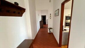 Un couloir avec un sol carrelé, un banc en bois, une porte donnant sur une cuisine, et un vestibule avec un radiateur et un cadre de tableau.