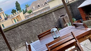 Une table et des chaises en bois sont disposées à l’extérieur, offrant une vue sur le paysage urbain et un mur de pierres.