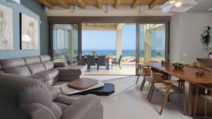 Een moderne woonkamer met een ruime hoekbank, een houten eettafel en stoelen. Grote ramen bieden uitzicht op de oceaan.