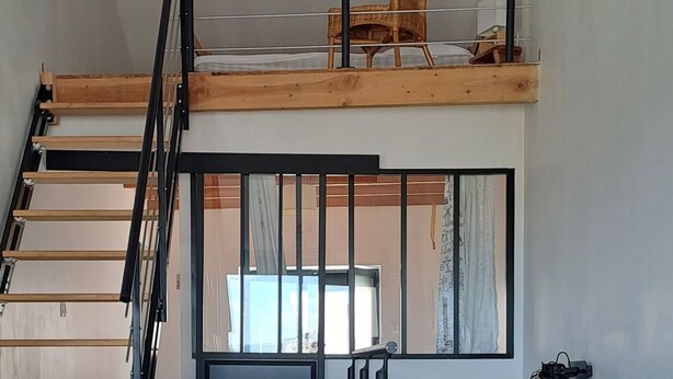 Ein Loft-Wohnbereich mit einer Couch, Kissen, einer Glasvase mit Weidenähren, einem Holzstuhl und einer Treppe.
