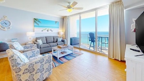 Ein geräumiges Wohnzimmer mit einer Couch, Sesseln, einem Couchtisch und einem Fernseher. Ein Balkon bietet Blick aufs Meer.