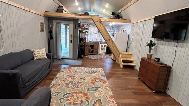 Un salon chaleureux avec un escalier en bois, un téléviseur à écran plat et un coin cuisine à l’arrière-plan.
