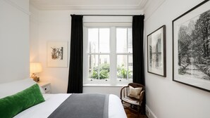 Ein Hotelzimmer mit einem Bett, einem Sessel, einem Nachttisch, einer Lampe, einem Fenster mit Vorhängen und gerahmten Bildern an der Wand.