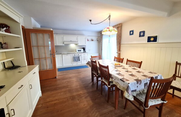 A dining area with a wooden table and chairs, a kitchen in the background, and a door leading to another room.
