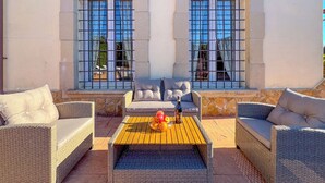 Un área de estar al aire libre iluminada por el sol, con muebles de mimbre y una mesa, dos ventanas con rejas de metal y una pared de piedra.
