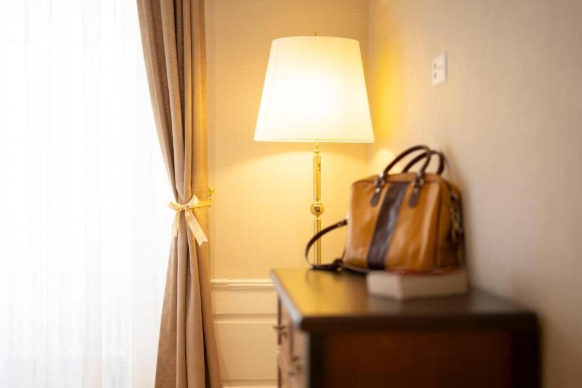 Ein Zimmer mit einer Lampe auf einem Nachttisch, einer Handtasche auf dem Tisch und einem zurückgebundenen Vorhang.