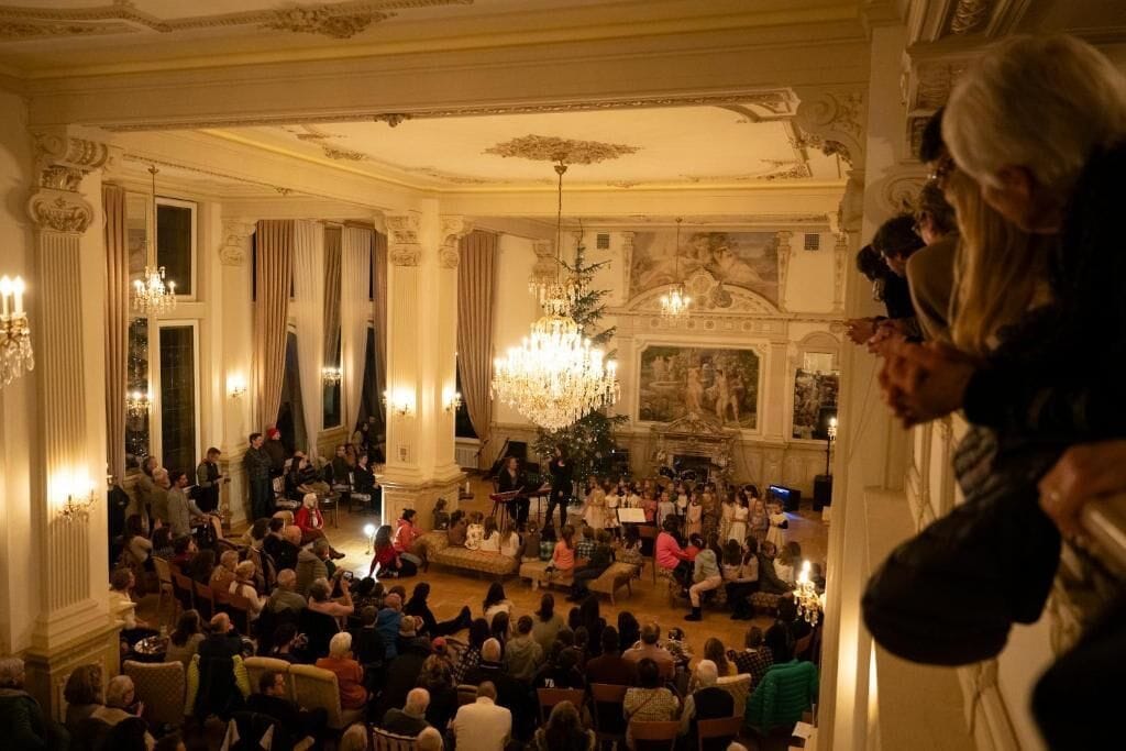 A grand hall with ornate columns, chandeliers, and a seated audience.