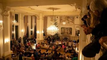 A grand hall with ornate columns, chandeliers, and a seated audience.