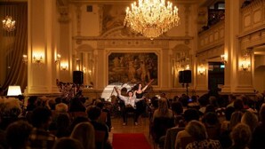 A grand hall with a red carpet, ornate chandeliers, and a large audience.