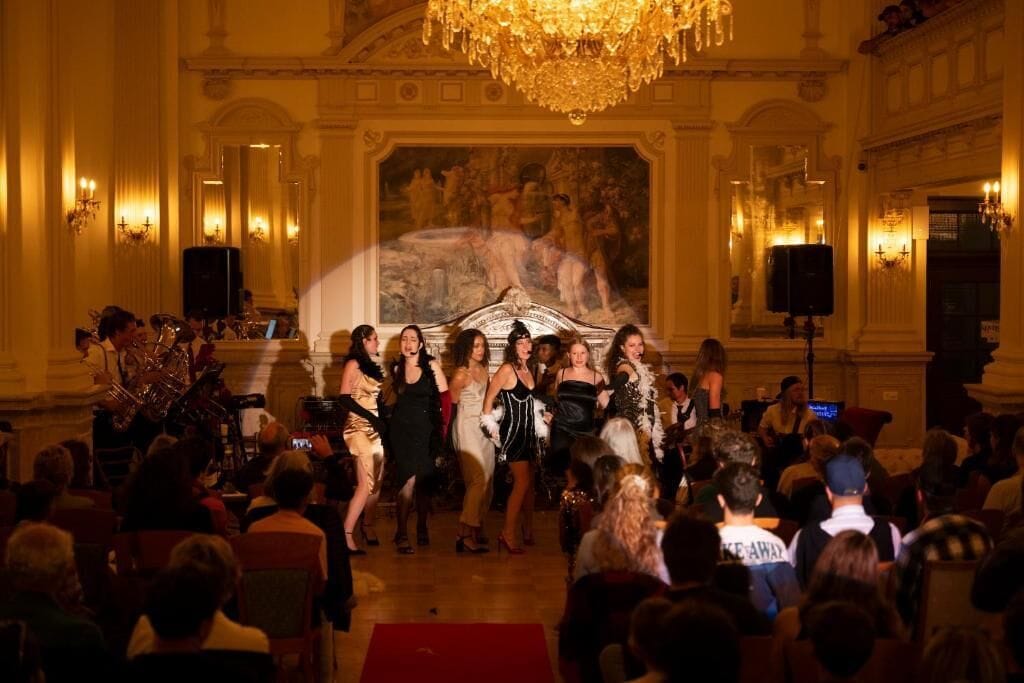 A formal event in a grand hall with a large audience, a stage with a classical painting, and ornate chandeliers.