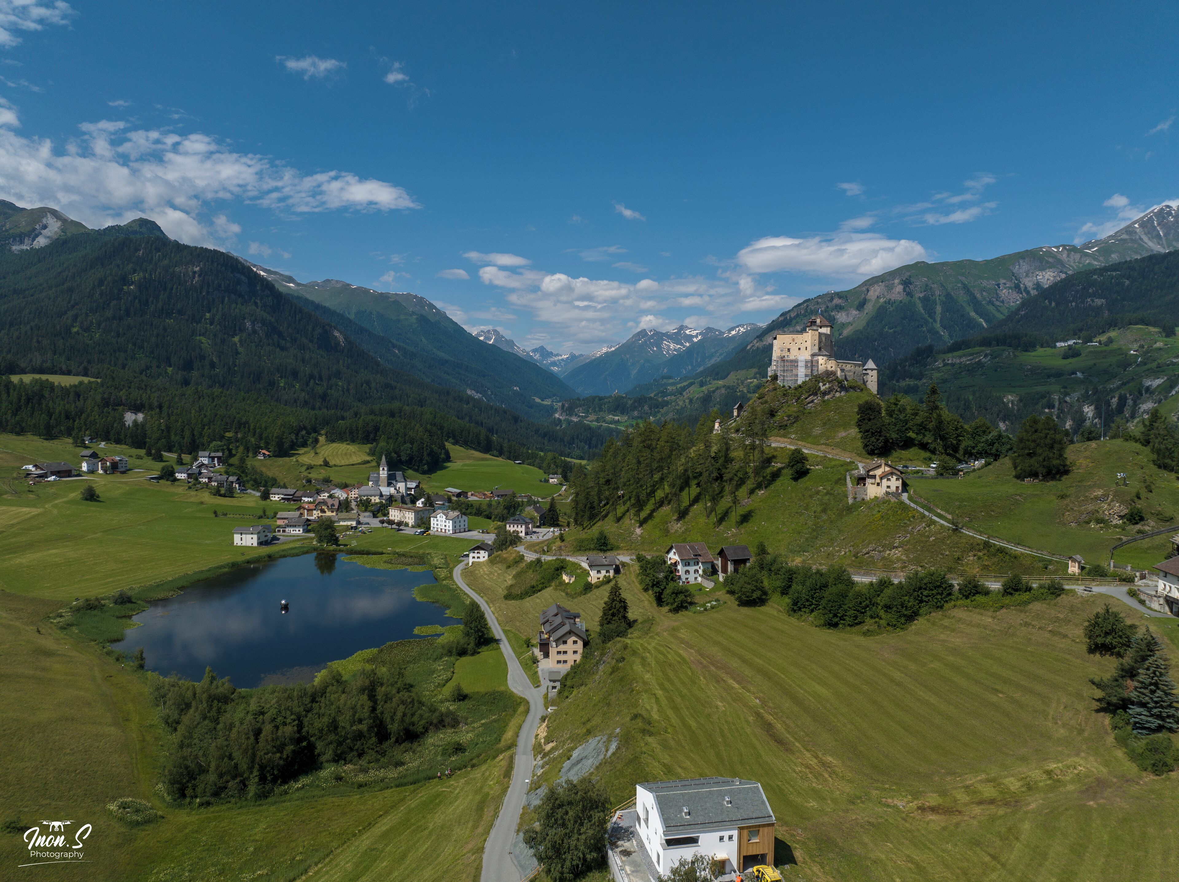 A castle on a hill overlooking a valley with a lake, surrounded by houses and greenery.