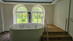A spa room with a large bathtub, a wooden staircase, and a view of a lush garden through arched windows.