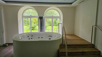 Un cuarto de spa con una bañera grande, una escalera de madera y vistas a un jardín exuberante a través de ventanas de arco.