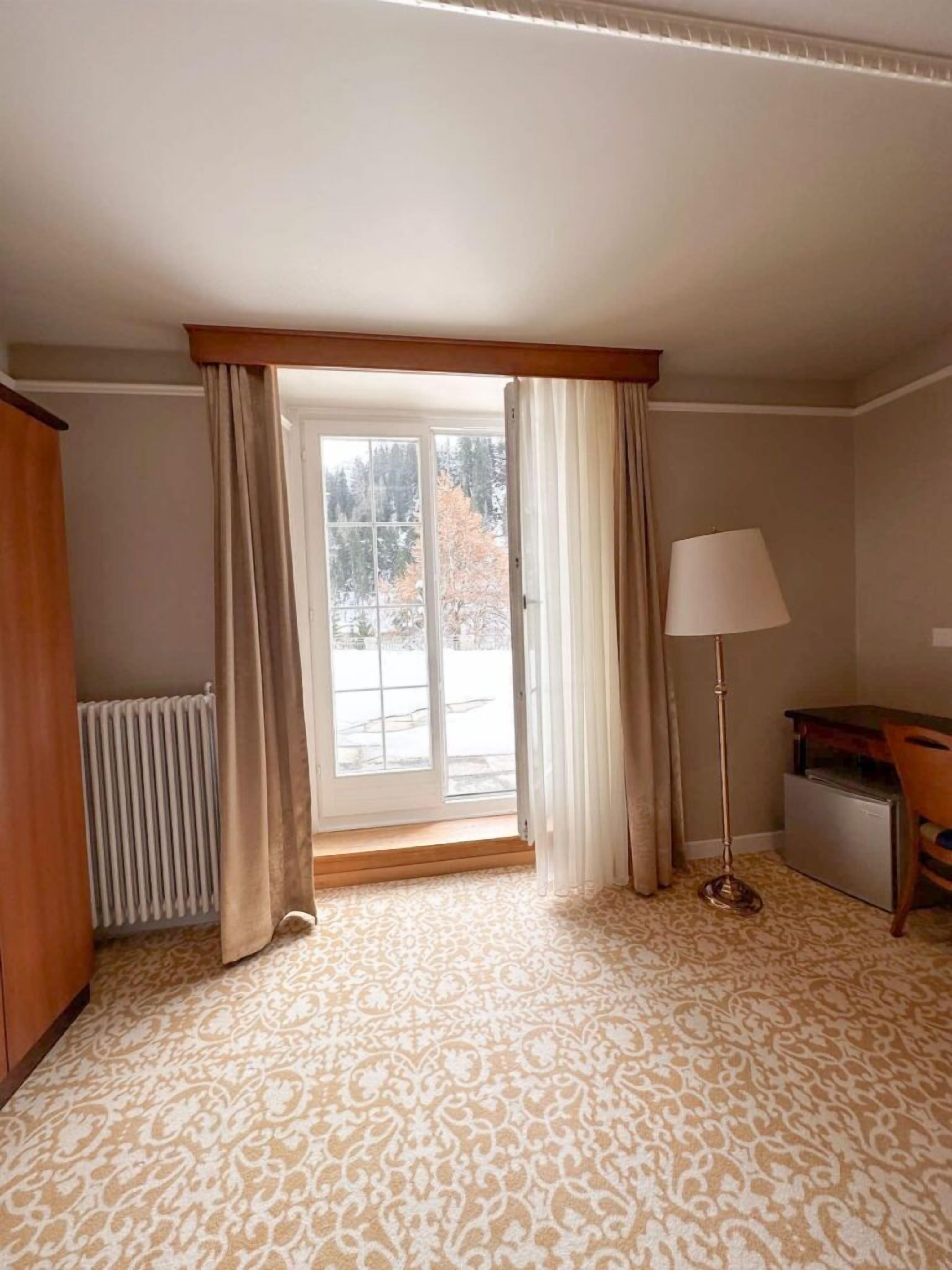 Ein Hotelzimmer mit einem gemusterten Teppich, einem Heizkörper, einem Schreibtisch mit Stuhl, einer Stehlampe und einem Fenster mit Vorhängen.