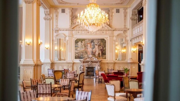 A grand hall with ornate columns, a chandelier, and a fireplace.