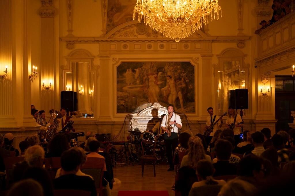 Een statige balzaal met gedetailleerde kroonluchters, een podium met muzikanten en een groot publiek.