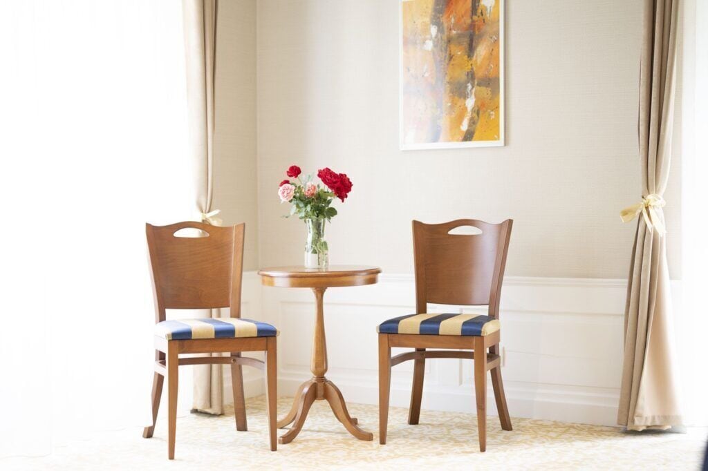 A room with two wooden chairs with blue and beige cushions, a small round wooden table with a vase of red and pink flowers, and a large abstract painting on the wall.