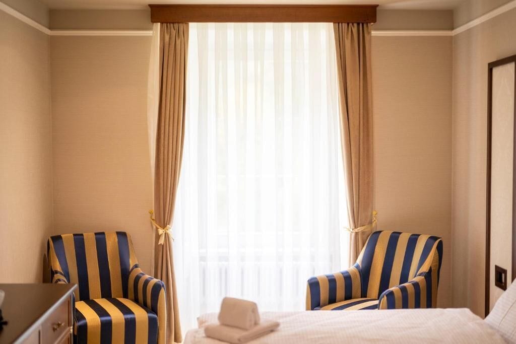Ein Hotelzimmer mit einem Bett, zwei gestreifte Ohrensessel, einem kleinen Tisch und einem Fenster mit leichtem Vorhang.