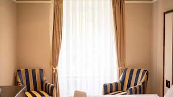 Ein Hotelzimmer mit einem Bett, zwei gestreifte Ohrensessel, einem kleinen Tisch und einem Fenster mit leichtem Vorhang.