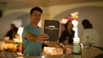 Una persona sosteniendo un libro titulado "SCUOL PALACE" en un bar, con una vela y vasos sobre la barra.