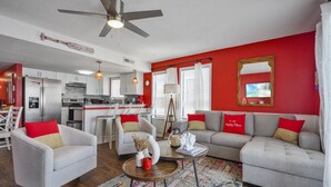 A modern living room with a red accent wall, a ceiling fan, a sofa set, and a kitchen in the background. - Sea Lion`s Den - Ocean View Condo, Steps to Sand (Carolina Beach)