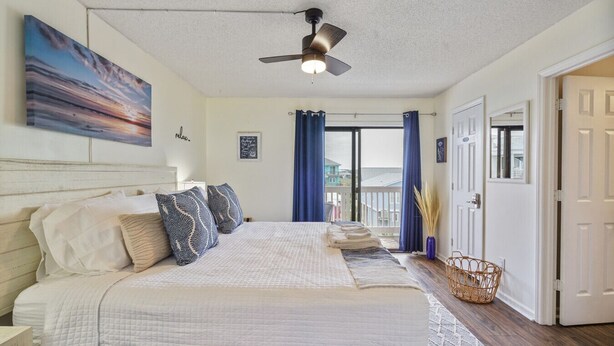 A bedroom with a large bed, a ceiling fan, a view to the outside, and a door to the right. - Sea Lion`s Den - Ocean View Condo, Steps to Sand (Carolina Beach)