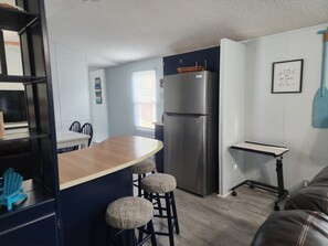 A compact kitchen with a stainless steel refrigerator, a wooden bar counter with stools, and a small table. - The Pelican's Nest — Cozy 3BR/2BA Oak Island Retreat, Sleeps 6 (Oak Island)