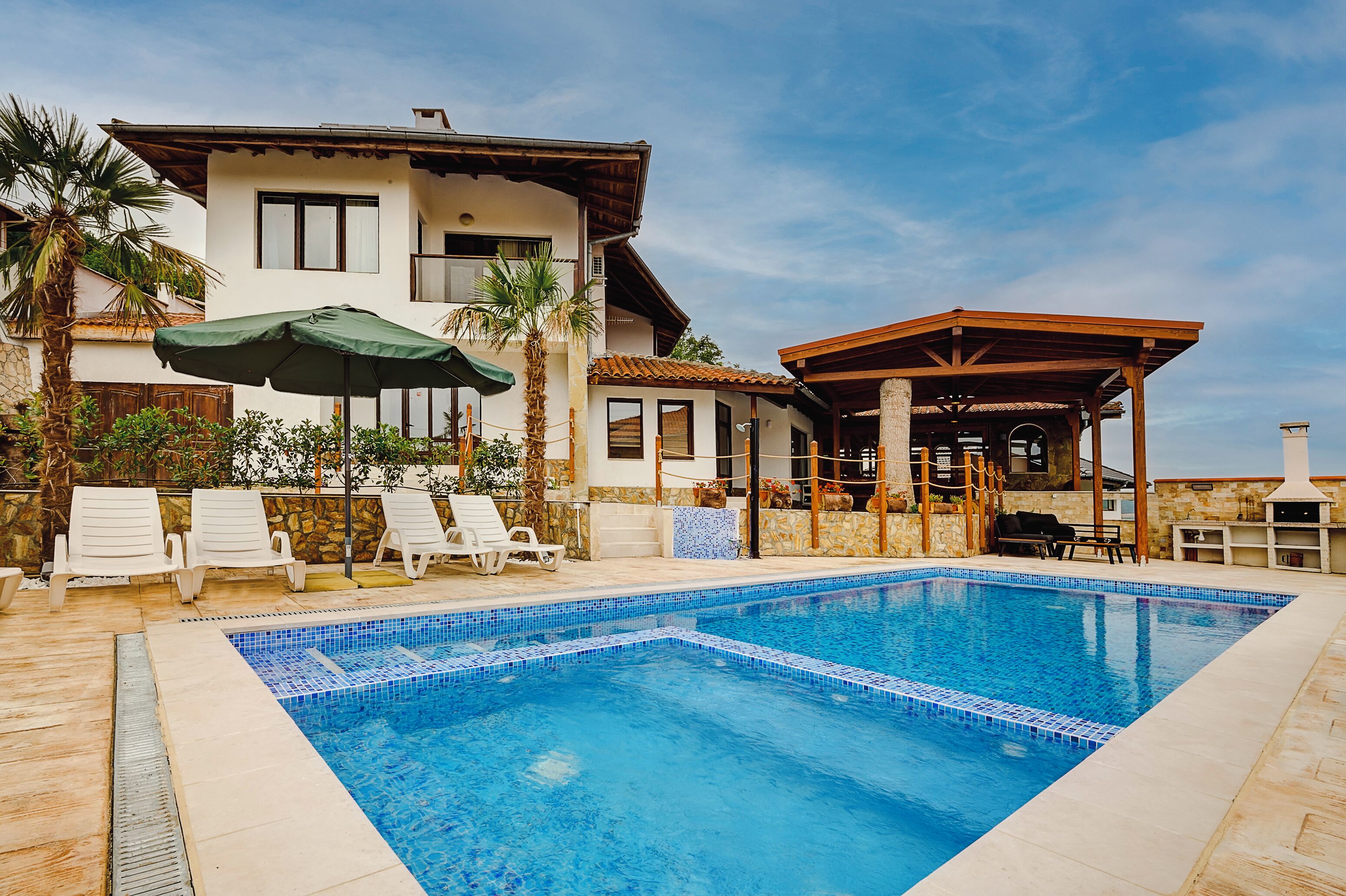 A swimming pool with lounge chairs and umbrellas, a covered patio, and a two-story house with a stone facade.