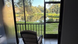A room with a sliding glass door leading to a balcony with a chair and a view of a pond and trees.