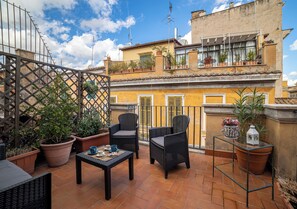 A terrace with wicker chairs and a glass table, surrounded by potted plants and a metal railing. - Palatino Rooftop Trastevere, a centrally located penthouse in Rome's Trastevere neighborhood, accommodating up to 6 people (Roma)