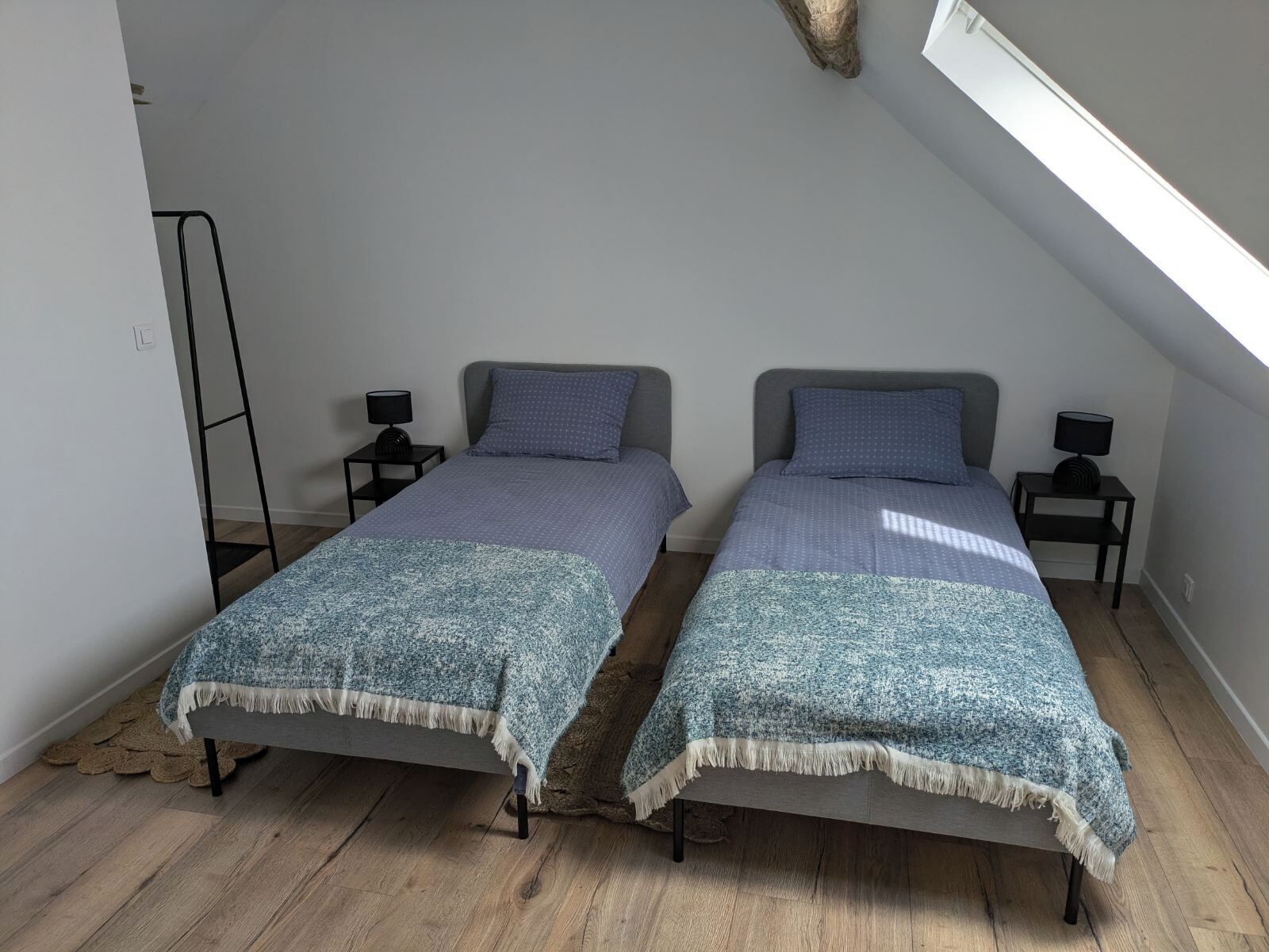 Une petite chambre moderne avec deux lits simples, chacun accompagné d’une table de chevet et d’une lampe assorties. La chambre est dotée d’un plafond en pente avec un puits de lumière, d’un plancher en bois et d’un aménagement simple et épuré.