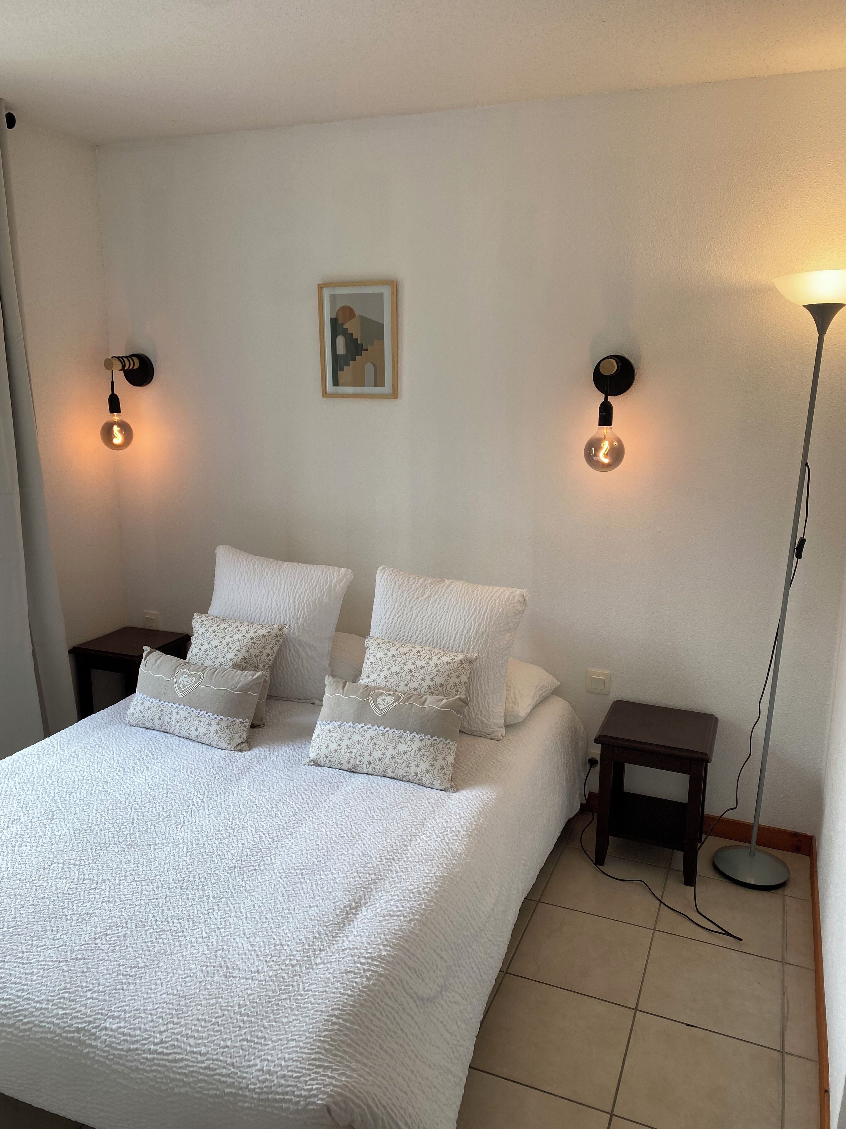 Un lit bien fait, avec une literie blanche et des coussins décoratifs, deux tables de chevet avec des lampes, un petit tableau accroché au mur et une lampe sur pied.