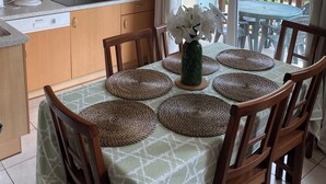 Un coin repas avec une table dressée pour quatre personnes, entourée de chaises en bois, d’un vase de fleurs blanches et de sets de table en fibres tressées.