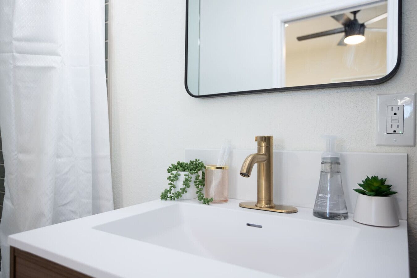 A bathroom with a white sink, a gold faucet, a mirror, a ceiling fan, and a potted plant.