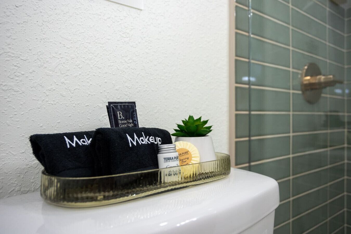 Ein Badezimmer mit einer weißen Toilette, einer Glasablage mit einem schwarzen Handtuch mit der Aufschrift 'Makeup', einer kleinen Topfpflanze und einer Dose Terra Pure Körperwaschgel.