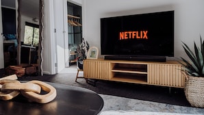 Ein Wohnzimmer mit einem großen Flachbildfernseher, der "NETFLIX" anzeigt, einer Holz-Unterhaltungsecke, einem Spiegel und einem Blumentopf.