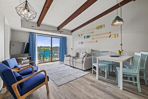 Ein modernes Wohnzimmer mit einer Couch, Sesseln, einem Esstisch und einem Fernseher. Holzdeckenbalken und ein Blick ins Freie durch eine Glastür.