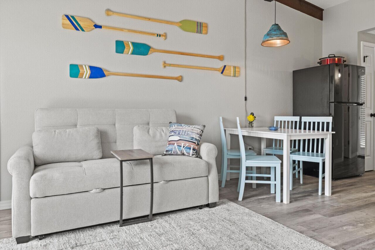 A living room with a grey sofa, a small table, and a dining area with a white table and blue chairs. There are wooden paddles on the wall and a modern kitchen with a refrigerator.