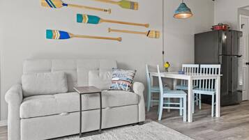 A living room with a grey sofa, a small table, and a dining area with a white table and blue chairs. There are wooden paddles on the wall and a modern kitchen with a refrigerator.