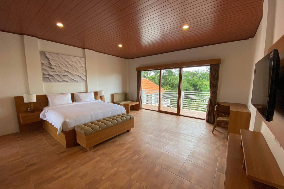 A spacious bedroom with a large bed, wooden headboard, bedside tables, a bench, a desk, and a TV.
