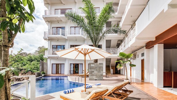 Zona de piscina con sillones de descanso y una sombrilla grande, rodeada por un edificio de varios pisos con balcones.