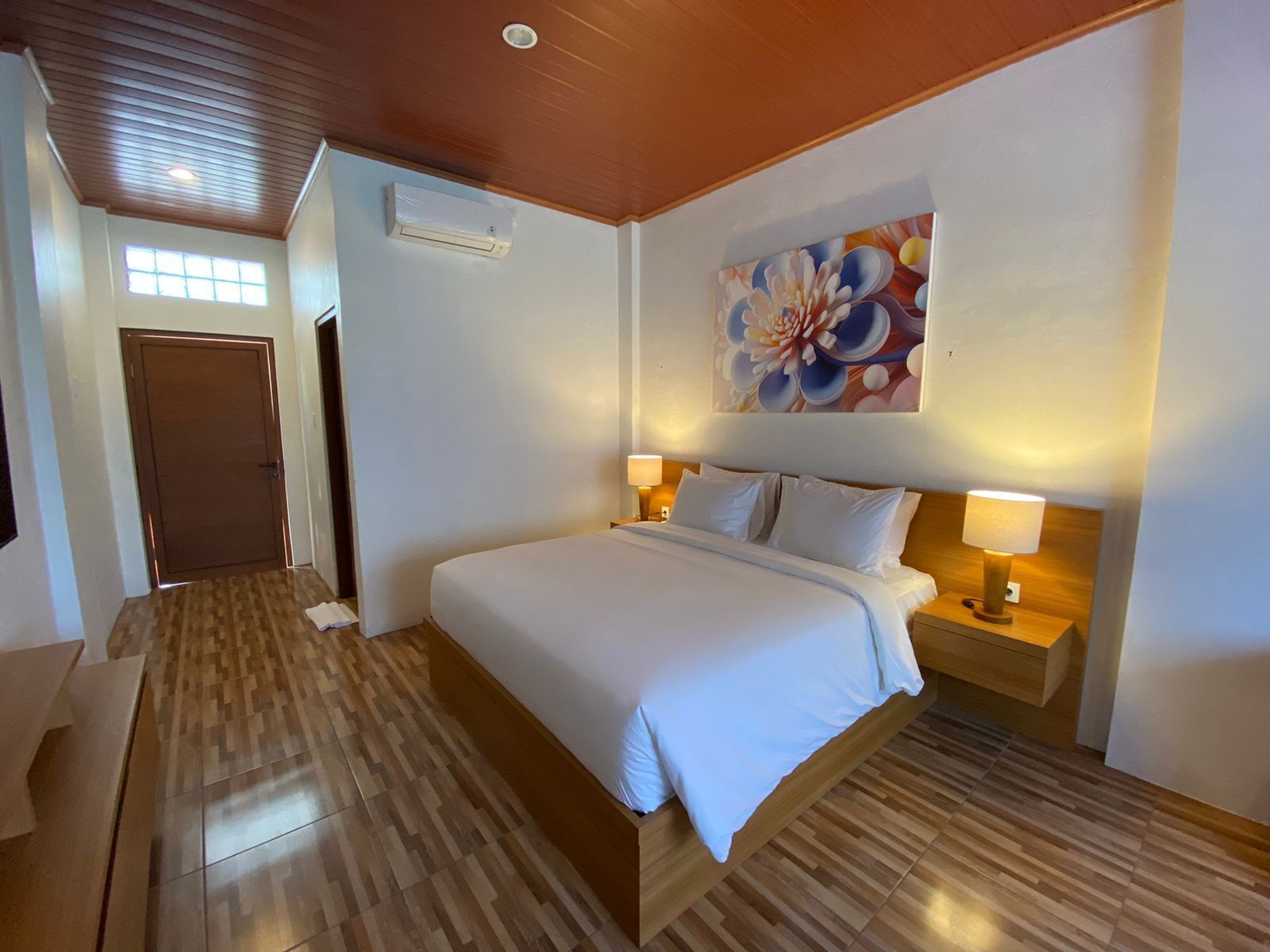A hotel room with a wooden floor, a bed with white linens, two bedside tables with lamps, a large floral artwork on the wall, and an air conditioning unit.