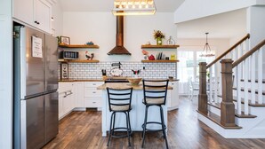 A kitchen with white cabinets, a stainless steel refrigerator, wooden bar stools, and a hanging light fixture. - Azur Hillside Haven: A Perfect Wellsboro Getaway! (Charleston Township)