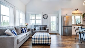 A spacious living room with a gray sofa, ottoman, and a kitchen area with a refrigerator and cabinets. - Azur Hillside Haven: A Perfect Wellsboro Getaway! (Charleston Township)